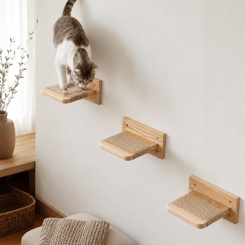 May include: A tabby cat explores a set of three wooden wall-mounted cat shelves. Each shelf has a natural fiber scratching surface. The shelves are staggered on a white wall, creating a climbing structure for feline enrichment.