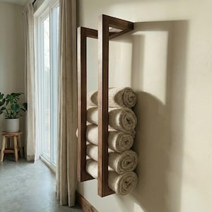 May include: A wooden towel rack mounted on a wall, holding rolled-up beige towels. The rack has a geometric design with vertical and horizontal wooden beams. The towels are neatly stacked, creating a clean and organized look. The rack is positioned near a window with natural light.