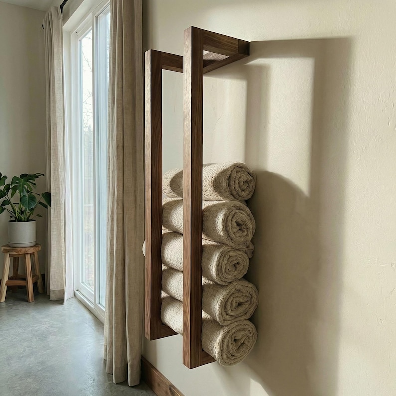 May include: A wooden towel rack mounted on a wall, holding rolled-up beige towels. The rack has a geometric design with vertical and horizontal wooden beams. The towels are neatly stacked, creating a clean and organized look. The rack is positioned near a window with natural light.