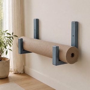May include: A wall-mounted yoga mat rack made of blue-stained wood with two L-shaped brackets. The rack holds a single rolled-up beige yoga mat. The brackets feature vertical mounting plates with visible screw holes for attachment to a flat wall surface.