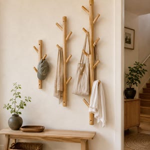 May include: Wooden wall-mounted coat racks in a tree branch design. A hat, scarf, tote bag, and towel hang from the branches. A wooden bench, basket, and vase with greenery are in the foreground. The room has a neutral colour palette.