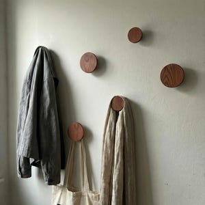 May include: Circular wooden wall hooks in a natural wood finish are mounted on a pale green wall. A grey jacket, a beige scarf, and a canvas tote bag hang from the hooks. The hooks have a minimalist design.