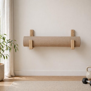 Op de afbeelding: Een opgerolde beige yogamat is aan een muur bevestigd met twee houten beugels. De mat wordt horizontaal gehouden en toont zijn textuur. De beugels zijn gemaakt van lichtgekleurd hout en bieden een minimalistisch en functioneel ontwerp voor fitness thuis.