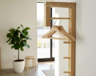 May include: A wooden wall-mounted coat rack with a rectangular frame and multiple pegs. Two wooden hangers hang from a horizontal bar. The rack is made of light-colored wood and is mounted on a white wall.
