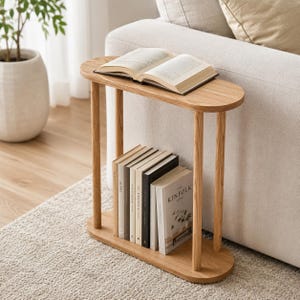 May include: A light-colored wooden side table with an oval top and bottom shelf, connected by four cylindrical legs. Several books are neatly arranged on the bottom shelf, and an open book rests on the top. The table is positioned next to a beige sofa.