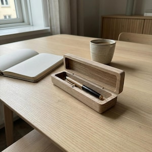 Pode incluir: Um estojo de caneta de madeira com uma caneta dourada e preta dentro, em cima de uma mesa de madeira clara. Um caderno e uma caneca de cerâmica também estão na mesa. O estojo da caneta está aberto, revelando a ponta da caneta.
