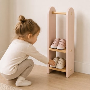 Könnte beinhalten: Ein rosa Schuhregal aus Holz mit zwei Regalen, auf denen Kinderschuhe stehen. Das Regal hat oben eine helle Holzstange und goldfarbene Akzente. Die Schuhe sind in Rosa- und Beigetönen gehalten. Ein Kind greift nach einem Paar Schuhe.