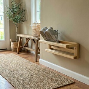 May include: A wooden shoe rack, holding a pair of grey trainers, is mounted on a beige wall. A woven jute rug lies on the wooden floor, leading to a wooden bench and a potted plant. A woven basket sits on the bench.