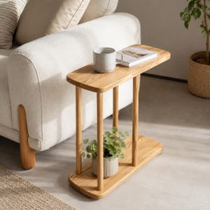 Op de afbeelding: Een lichte eikenhouten bijzettafel met een rechthoekig blad en een onderste plank. Op de plank staat een kleine plant in een pot. Een mok en een boek liggen op het tafelblad. De tafel staat naast een crèmekleurige bank.