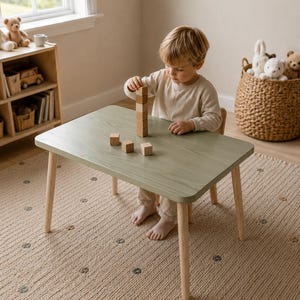 以下が含まれることがあります： 明るい色の脚が付いた、セージグリーンの小さな木製テーブル。子供がテーブルの上で木製のブロックで遊んでいます。テーブルは、窓、本棚、ぬいぐるみでいっぱいのバスケットのある部屋にあります。