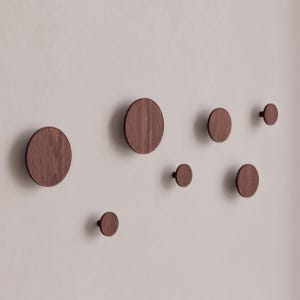 Op de afbeelding: Zeven ronde houten wandhaken in verschillende maten, allemaal in een donkerbruine houtkleur. De haken zijn gemonteerd op een witte muur.