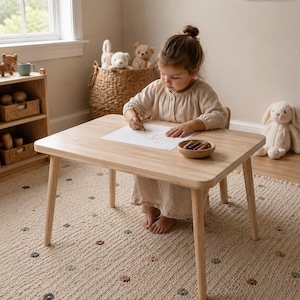 Op de afbeelding: Een kindertafel van licht hout met afgeronde hoeken en poten. Een klein kind zit aan de tafel en tekent met kleurpotloden op een stuk papier. Een kleine houten kom met kleurpotloden staat op de tafel. De kamer heeft een neutraal kleurenpalet.
