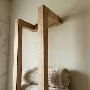 May include: A light-colored wooden towel rack mounted on a wall. The rack has a rectangular frame with two vertical supports and a horizontal top section. Rolled beige towels are visible on the bottom of the rack. The wood grain is visible.