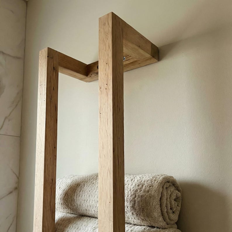 May include: A light-colored wooden towel rack mounted on a wall. The rack has a rectangular frame with two vertical supports and a horizontal top section. Rolled beige towels are visible on the bottom of the rack. The wood grain is visible.