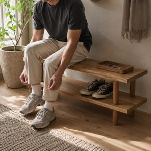 May include: A two-tiered wooden bench with a person sitting on it, tying their shoelaces. The bench has a natural wood finish. Two pairs of sneakers are on the bench. A small wooden tray with keys is on the top shelf.