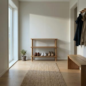 Può includere: Un ingresso luminoso con un porta scarpe, una panca e un attaccapanni in legno. Un tappeto tessuto è sul pavimento in legno. Le scarpe sono disposte sul porta scarpe. Una piccola pianta in vaso è vicino a una grande finestra. I cappotti sono appesi al muro.