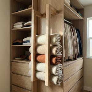 May include: A wooden closet with shelves and drawers. Rolled blankets in various neutral colors are displayed on a wooden rack. Clothes hang on a rod to the right, and folded clothes are on the shelves.