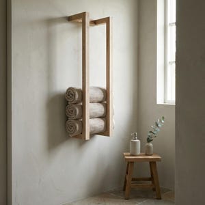 May include: A light-colored wooden towel rack mounted on a wall, holding rolled beige towels. The rack has a rectangular design. A small wooden stool with a soap dispenser and vase is next to the rack.