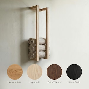 May include: A wooden towel rack with three rolled towels. The rack is made of light-colored wood and is mounted on a wall. The image also shows color options: Natural Oak, Light Ash, Dark Walnut, and Black Stain.