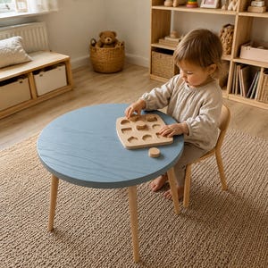 May include: A light blue round wooden table with light wood legs. A wooden puzzle with geometric shapes sits on the table. A child is seated at the table, playing with the puzzle. The table is approximately 24 inches in diameter.