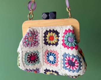 Puede incluir: Un bolso de mano de crochet blanco con un marco de madera y un estampado floral colorido. El bolso tiene una correa de cadena dorada y morada.