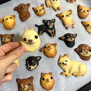 Puede incluir: Una bandeja para hornear con una variedad de galletas con forma de perro. Las galletas están decoradas con diferentes colores y diseños, incluyendo marrón, negro, blanco y amarillo. Algunas de las galletas tienen ojos saltones y otras tienen detalles pintados. Las galletas están dispuestas en un patrón aleatorio en la bandeja para hornear. El texto '@Yirannnn' es visible en la bandeja para hornear.