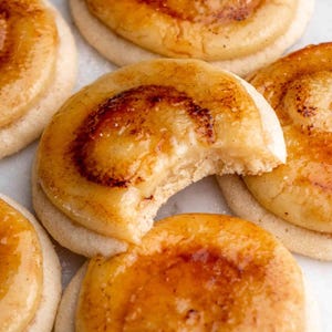 May include: Close-up of a group of round cookies with a caramelised sugar topping. The cookies are arranged on a white surface. One cookie has a bite taken out of it, revealing a creamy filling.