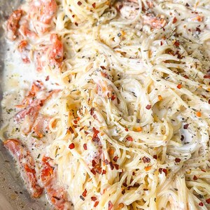 Puede incluir: Primer plano de un plato de pasta cremosa con espaguetis, tomates rojos y hierbas. La pasta est&aacute; cubierta con una salsa blanca y espolvoreada con copos de pimiento rojo. Una comida deliciosa y apetitosa.