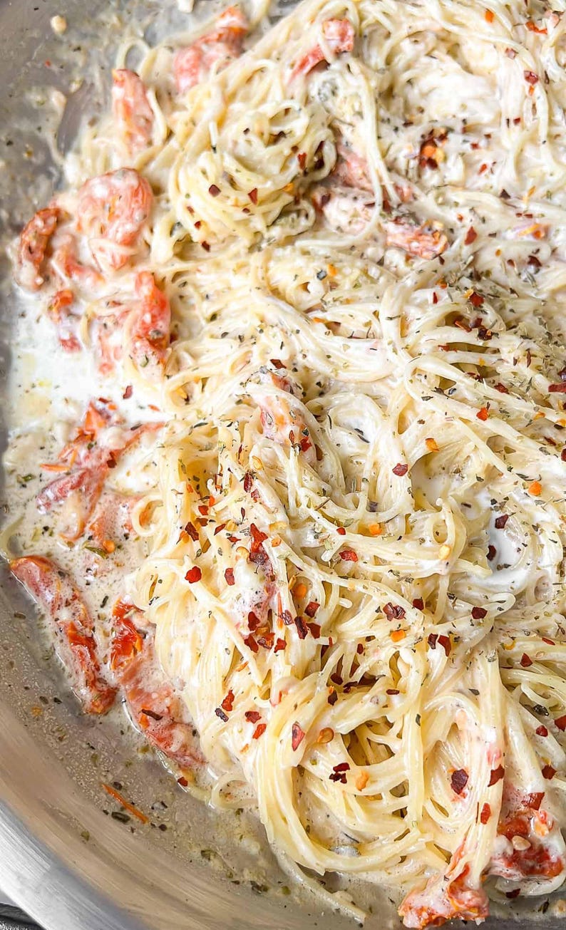 Puede incluir: Primer plano de un plato de pasta cremosa con espaguetis, tomates rojos y hierbas. La pasta est&aacute; cubierta con una salsa blanca y espolvoreada con copos de pimiento rojo. Una comida deliciosa y apetitosa.