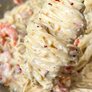 Puede incluir: Primer plano de un plato de pasta cremosa con ingredientes rojos y verdes. La pasta est&aacute; sujeta por pinzas plateadas, mostrando la salsa cremosa y los ingredientes. El plato est&aacute; en una sart&eacute;n plateada.