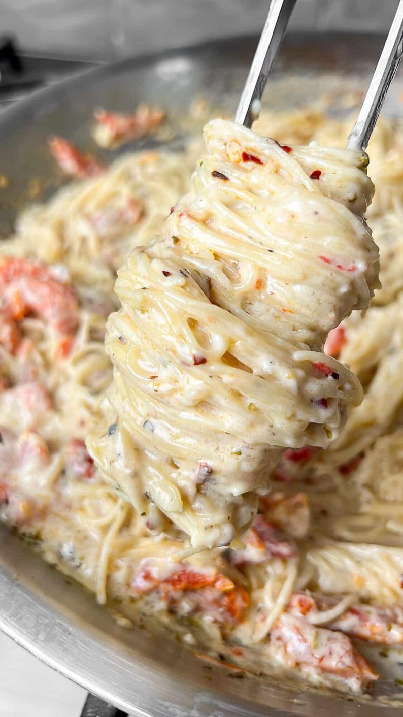 Puede incluir: Primer plano de un plato de pasta cremosa con ingredientes rojos y verdes. La pasta est&aacute; sujeta por pinzas plateadas, mostrando la salsa cremosa y los ingredientes. El plato est&aacute; en una sart&eacute;n plateada.