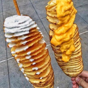 Op de afbeelding: Twee spiraalvormige frietjes op stokjes, een met witte saus en de andere met gesmolten kaas.