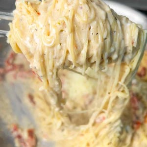 Puede incluir: Primer plano de espaguetis con salsa cremosa blanca y tomates rojos. La pasta se levanta de una sart&eacute;n de acero inoxidable, mostrando la textura y los ingredientes. Un plato delicioso.