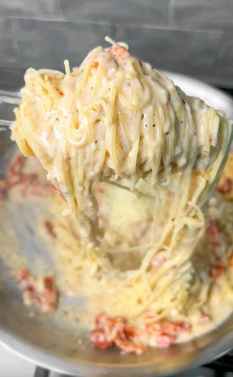 Puede incluir: Primer plano de espaguetis con salsa cremosa blanca y tomates rojos. La pasta se levanta de una sart&eacute;n de acero inoxidable, mostrando la textura y los ingredientes. Un plato delicioso.
