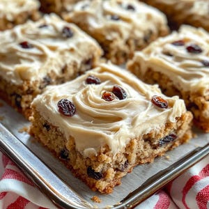 May include: Close-up of several frosted raisin bars on a metal baking sheet. The bars have a light brown base with dark raisins and are topped with a swirl of creamy frosting and more raisins. A red and white checkered cloth is visible.