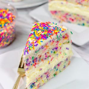 Puede incluir: Una rebanada de pastel con glaseado blanco y chispas de colores. El pastel está en capas y tiene un relleno blanco. El pastel está en un plato blanco con un tenedor dorado.
