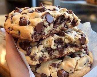 May include: Three freshly baked chocolate chip cookies stacked on top of each other, with a light brown exterior and a soft, chocolate-filled interior. The cookies are held together by a white paper.