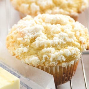 May include: Close-up of a golden-brown muffin with a crumbly, white streusel topping. The muffin sits in a paper liner, with a partially visible stick of butter in the foreground. The baked good is on a metal cooling rack.