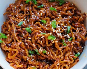 May include: A white bowl filled with spicy noodles topped with sesame seeds and green onions.
