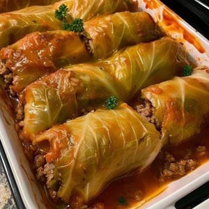 Op de afbeelding: Een close-up van verschillende koolrolletjes in een witte ovenschaal. De koolbladeren zijn lichtgroen en de vulling is een mengsel van vlees en rijst. De rollen zijn bedekt met een rode saus en gegarneerd met peterselie.