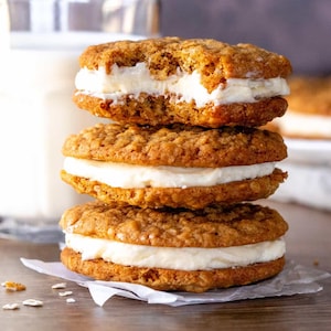 Puede incluir: Una pila de tres galletas de avena con un relleno blanco cremoso. Las galletas están sobre un trozo de papel de pergamino y hay un vaso de leche en el fondo.