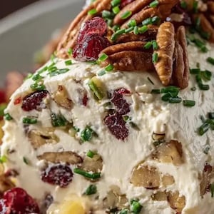 Puede incluir: Primer plano de un tronco de queso cremoso y blanco cubierto con nueces pecanas, arándanos rojos secos y cebollino fresco. El tronco está salpicado de nueces y pistachos.