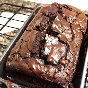 May include: A close-up of a chocolate loaf cake with a rich, dark chocolate glaze. The cake is cracked and has a slightly uneven surface, indicating it is homemade. The cake is in a dark gray rectangular baking pan.