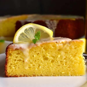 Könnte beinhalten: Ein Stück Zitronenkuchen mit einem Zitronenspalten auf der Oberseite und einem weißen Guss darüber. Der Kuchen liegt auf einem weißen Teller.