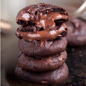 May include: A stack of four dark chocolate cookies, one cut open to reveal a rich, flowing chocolate filling. The cookies are a deep brown colour, with a slightly textured surface. A sprinkle of sea salt is visible on top.