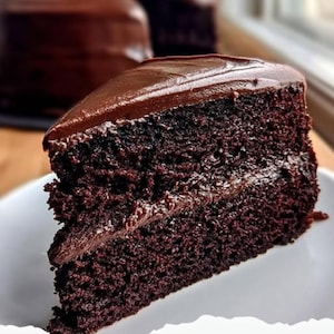 以下が含まれることがあります： 白いお皿にのった、濃厚なチョコレートケーキのスライス。ケーキは、ダークチョコレートのフロスティングが塗られており、しっとりとした質感で、ケーキの層の間にはフロスティングの層があります。背景には別のケーキが見えます。