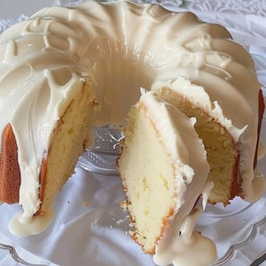 Puede incluir: Un bizcocho bundt en rodajas con un glaseado blanco cremoso. El bizcocho es de color amarillo claro con una corteza dorada. Se ha retirado una rebanada, revelando la textura del bizcocho. El glaseado gotea por los lados, creando un postre visualmente atractivo.