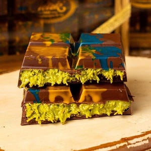 May include: Two stacked chocolate bars with a pistachio filling. The dark chocolate exterior is decorated with streaks of yellow and blue paint. The filling is a bright green color, creating a striking contrast.