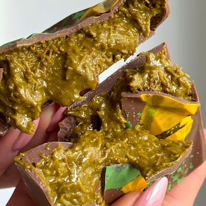 Op de afbeelding: Close-up van een in tweeën gebroken chocoladereep, met een groene vulling. De chocolade is donkerbruin met groene en gele accenten. De vulling heeft een gescheurde textuur en de reep wordt door een persoon vastgehouden.