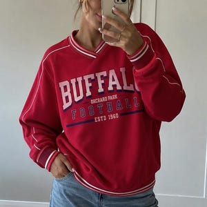 May include: A red long-sleeve sweatshirt with a V-neck collar and white and red striped trim. The front features white text reading BUFFALO, with smaller text reading ORCHARD PARK, FOOTBALL, and ESTD 1960. Paired with light blue wide-leg denim jeans.